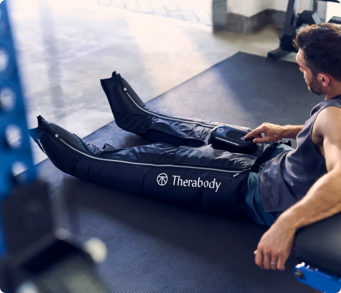 A man in a sleeveless shirt lies on a mat, using Therabody recovery boots on his legs, as he looks at a device or controls for the boots.